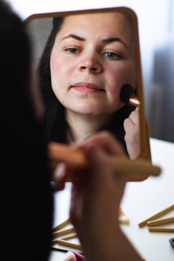 Millennial Kafkas kadınının makyaj tozu sürerken aynaya baktığı dikey görüntüsü. Aynadaki yansıma. Hazırlanıyor. Güzellik konsepti.