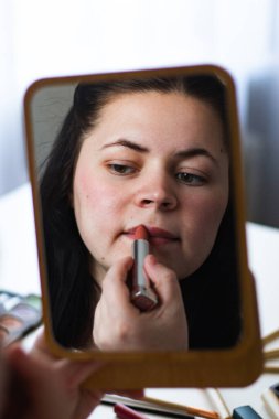 Bin yıllık beyaz bir kadının aynaya bakarken dudaklarına ruj sürerek makyaj yaptığı dikey bir görüntü. Aynadaki yansıma. Hazırlanıyor. Güzellik konsepti.