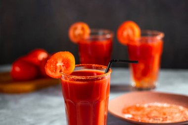 Bardakta üç Bloody Mary kokteyli ve gri taşlı yüzeyde domates dilimi. Kırmızı kokteyl. Alternatif tat. Kirli içki.