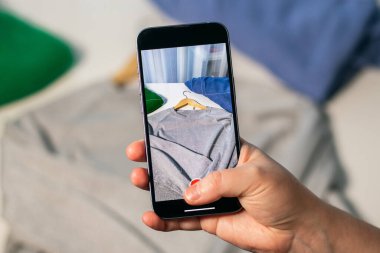 Akıllı telefonlu bir kadın kullanılmış gömleğin fotoğrafını çekiyor. İkinci el çevrimiçi dükkanda satılacak kıyafetleri hazırlıyorum. Malları ve ikinci el konsepti sat. Kıyafet bağışı