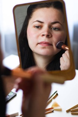 Millennial Kafkas kadınının makyaj tozu sürerken aynaya baktığı dikey görüntüsü. Aynadaki yansıma. Hazırlanıyor. Güzellik konsepti.