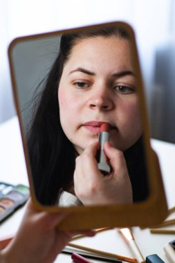 Bin yıllık beyaz bir kadının aynaya bakarken dudaklarına ruj sürerek makyaj yaptığı dikey bir görüntü. Aynadaki yansıma. Hazırlanıyor. Güzellik konsepti.