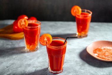 Bardakta üç Bloody Mary kokteyli ve gri taşlı yüzeyde domates dilimi. Kırmızı kokteyl. Alternatif tat. Kirli içki.