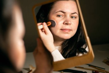 Evde makyaj yapan bir kadın aynaya bakıyor. Fırçayla makyaj yapıyorum. Yüzüne cilt pudrası sürüyorum. Kırmızı noktaları kaplıyor. Aynadaki yansıma.