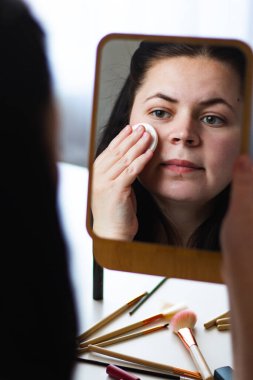 Millennial Kafkas kadınının aynaya bakarken yüzünden makyajı çekerken dikey görüntüsü. Aynadaki yansıma. Yatmaya hazırlanıyorum. Kişisel bakım güzellik konsepti.