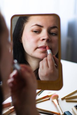 Bin yıllık beyaz bir kadının aynaya bakarken dudaklarına ruj sürerek makyaj yaptığı dikey bir görüntü. Aynadaki yansıma. Hazırlanıyor. Güzellik konsepti.