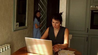 Genç anne, kızı koluna yaslanıp birlikte zaman geçirmek isterken mutfak masasında uzaktan çalışmayı yönetiyor. Görüntü, aile ortamında kariyer ve çocuk bakımı arasındaki farkı yansıtıyor