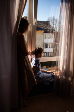 Anne kızının saçını örüyor, sevgi ve bağ anı yaratıyor. Çocukluk güzelliği, aile bağı ve hassas bakım. Ebeveynlik ve aile yaşam tarzı temaları için görüntü