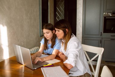 Anne ve kızı evde otururken dizüstü bilgisayar kullanıyorlar. Anne okul ödevlerinde çocuğa yardım ediyor. Modern teknolojilerle eğitim.