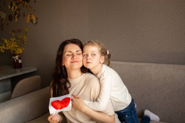 Açık renk saçlı genç kız çocuksu çizimini tutarken annesine sıkıca sarılıyor. Saf anne-kız aşkı.