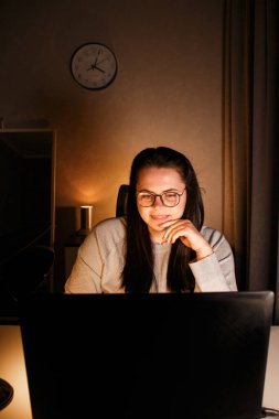 Gözlüklü genç bir kadın gece geç saatlerde evde bilgisayar başında çalışıyor, modern teknolojileri kullanıyor ve çevrimiçi eğitim alıyor. Uzaktan öğrenme konsepti. Online üniversite. Sürece odaklan.
