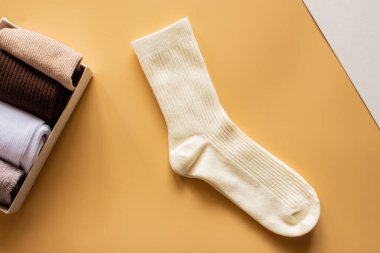 Sarı arka planda sergilenen bir çift krem renkli çorap, çeşitli renklerde katlanmış çorapların yanında, tekstil dokusu ve modern moda aksesuarları sergileniyor..