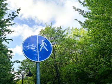 Parlak gökyüzü altında yemyeşil ağaçlarla çevrili bisiklet ve yayalar için ayrı yolları gösteren mavi dairesel trafik levhası, güvenli açık hava aktivitelerini ve şehir planlamasını teşvik ediyor..