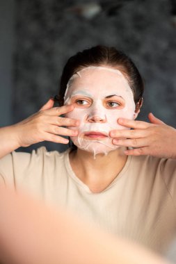 Banyodaki aynaya kumaş maske takan genç bir kadın. Cilt bakımı, kişisel bakım ve güzellik tedavi kavramları. Uluslararası Kişisel Bakım Günü