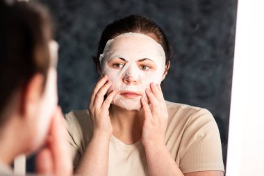 Geç saatte cilt bakımı rutini. Nemlendirici yüz maskesi takan bir kadın. Banyo aynasında yansıma. Dermatoloji kavramı