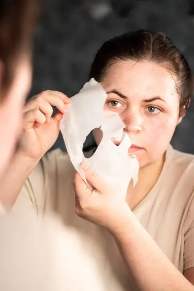 Banyo aynasında çarşaf maskesini çıkaran bir kadın taze, parlak bir cilt olduğunu gösteriyor. Bakım sonrası cilt bakımları ve güzellik ürünleri promosyonları için mükemmel.