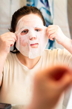 Banyodaki aynaya kumaş maske takan genç bir kadın. Cilt bakımı, kişisel bakım ve güzellik tedavi kavramları. Uluslararası Kişisel Bakım Günü