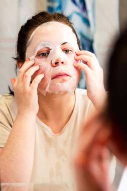 Akşam cilt bakımı rutini. Besleyici yüz maskesi takan bir kadın. Geç saatte yüz bakımı. Banyo aynasında yansıma. Dermatoloji kavramı