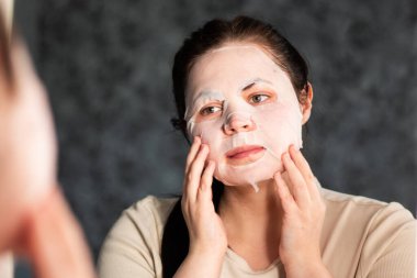 Akşam cilt bakımı rutini. Besleyici yüz maskesi takan bir kadın. Geç saatte yüz bakımı. Banyo aynasında yansıma. Dermatoloji kavramı