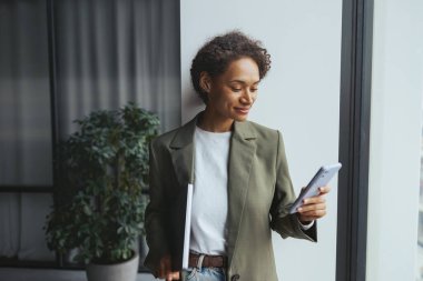 Resmi kıyafetli bir kadın elinde bavulla telefonunu kontrol ediyor. Kapının yanında duruyor, ceketinin kolları sarılmış, belini ve kalçasını bir çiçeğe yaslarken tek eliyle işaret ediyor.