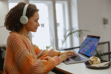 Kulaklıklı bir kadın masadaki dizüstü bilgisayarda yazıyor. Laptop, ofis malzemeleri ve ekipmanlarıyla çevrili bir masanın üzerine konur.