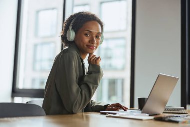 Bir beyaz işçi, bir kadın, tahta bir masada dizüstü bilgisayar, bir çıkış cihazı kullanarak otururken gülümsüyor. Pencerenin yanında.