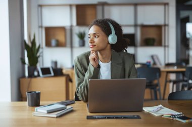 Kulaklıklı bir kadın bir masada oturuyor, binadaki dizüstü bilgisayarın karşısında. Masanın üzerinde saksıda bir saksı var.