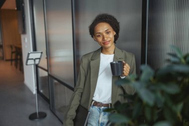 Resmi kıyafetli bir beyaz işçi koridorda elinde bir fincan kahve, gözlük ve kapının yanında bir bitki olan bir kolla durur.