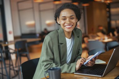 Beyazlar işçi kıyafeti giymiş bir kadın dizüstü bilgisayar ve cep telefonuyla bir masada oturuyor ve bilgisayarda çalışırken gülümsüyor.