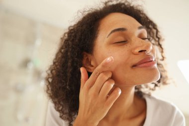 Bir kadın nazikçe alnına, burnuna, yanaklarına, dudaklarına ve boynuna losyon sürerken aynadaki yansımasına hayran oluyor ara sıra gülümsüyor ve kirpiklerini fırçalıyor.