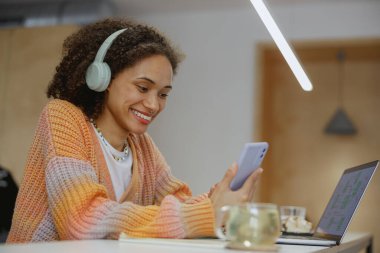 Kulaklık takan mutlu bir kadın, bir masada dizüstü bilgisayar ve cep telefonuyla oturuyor. Ses ekipmanı ve kişisel bilgisayar kullanırken gülümsüyor.