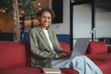 Bir kadın kırmızı bir kanepede oturuyor, dizüstü bilgisayar kullanıyor. Binanın iç tasarımı, o gülümserken rahatlık ve rahatlık sağlıyor.