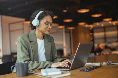 Kulaklıklı bir kadın masasında dizüstü bilgisayarında yazı yazıyor. Kullandığı elektronik cihaz kişisel bir bilgisayar. O da bir şapka ve ceket giyiyor.