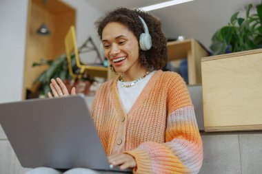 Bir kadın dizüstü bilgisayar kullanırken ve kulaklık takarken gülümsüyor. Onun yanındaki dolapta bir saksı bitkisi var. Elektronik cihazın teknolojisinden zevk alıyor.