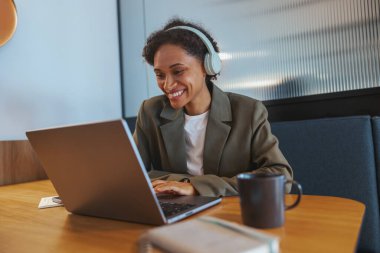 Bir beyaz işçi bir masada oturuyor, kulaklığı olan bir dizüstü bilgisayar kullanıyor. Ahşap bir masada kişisel bilgisayarı üzerinde çalışırken odaklanmış ve gülümsüyor.