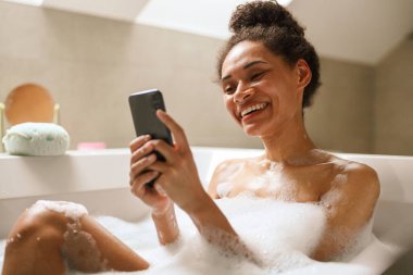 Resmi giyinmiş bir kadın telefonuna bakarken mutlu bir şekilde banyo yapıyor. Baş parmağıyla gülümsüyor ve jestler yapıyor, muhtemelen düğün organizasyonlarının detaylarını ya da düğün aksesuarlarını kontrol ediyor.