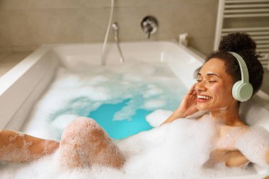 Bir kadın su dolu bir küvette mutlu bir şekilde banyo yapar, kulaklık takar, banyoda sakin ve eğlenceli bir anın tadını çıkarır, yüzünde bir gülümsemeyle.