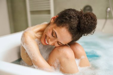 Bir kadın küvette banyo yapıyor, çıplak ve mutlu bir şekilde gülümsüyor. Göğüs ve kalçaları kısmen görülüyor. Banyodaki rahatlatıcı sıvıdan hoşlanıyor.