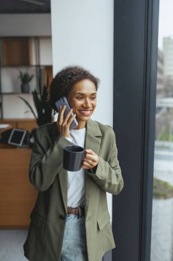 Resmi bir ceket ve takım elbise giyen kadın, bir elinde bir fincan kahve tutarken diğer eliyle cep telefonuna konuşurken gülümseyerek hareket eder.