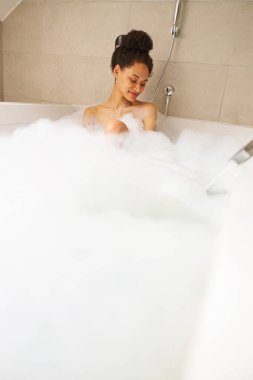 Bir kadın köpük dolu bir küvette banyo yapıyor. Kafası kenarda duruyor, yüzünü ve saçlarını baloncuklar kaplıyor. Boru tesisatıyla çevrili bir elbise giyiyor.