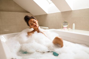 Bir kadın köpüklü sıvıyla dolu bir küvette rahatlatıcı bir banyonun keyfini çıkarıyor. Banyoda, malzeme döşemesi ve tesisat tesisatı olan bir yerde.