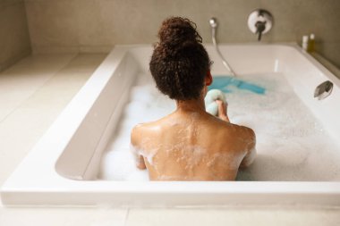 Bir kadın suyla dolu bir küvette banyo yapıyor. Temizlenmek için sünger kullanıyor. Su tesisatı ve küvetin bileşik malzemesi o banyonun tadını çıkarırken sıvıyı tutuyor.