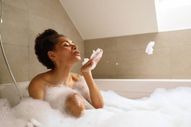 Çıplak bir jest yapan bir kadın küvette banyo yaparken sabun köpüğü üflüyor. Odanın parke döşemesi ve tavanı olaylara sakin bir ortam hazırladı.