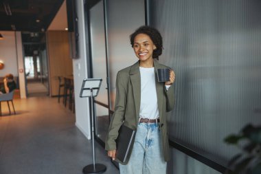 Bir kadın koridorda elinde bir fincan kahve ve dizüstü bilgisayarla dikiliyor, şık bir kolla resmi giysiler giyiyor, bir moda etkinliği sırasında binanın penceresinden dışarı bakıyor.