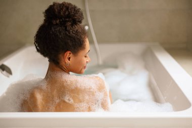 Bir kadın köpük ve suyla dolu bir küvette banyo yapıyor. Tesisat tesisatının sağladığı rahatlığın tadını çıkarıyor. Sıcak suda ıslanırken küvetin borusu açık.