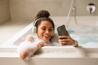 Bir kadın küvette gülümsüyor, taşınabilir bir iletişim cihazı kullanıyor ve cep telefonundan müzik dinlerken başparmağıyla işaret ediyor.