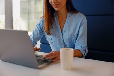 Mavi gömlekli bir kadın iyi aydınlatılmış bir ofiste elinde kahve fincanıyla dizüstü bilgisayarda yazı yazıyor. Parlak ve profesyonel görünüyor.