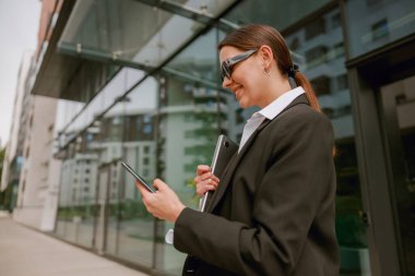 Profesyonel bir iş kadını, modern bir ofis binasının önünde dururken tablet kullanır ve belgeleri saklar.