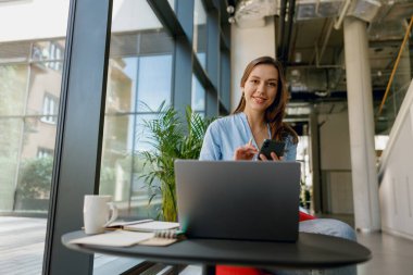 Genç profesyonel bir kadın, dizüstü bilgisayarı ve akıllı telefonuyla modern, aydınlık bir ofis alanında görevleri yönetiyor.