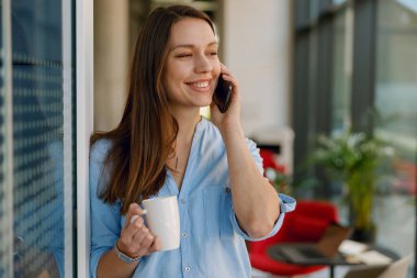 Neşeli bir kadın telefon görüşmesi yapıyor ve elinde parlak ve modern bir ofis ortamında kupa tutuyor..